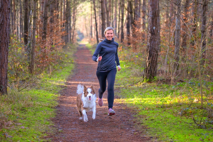 A middle-aged woman jogging with her dog on a trail.