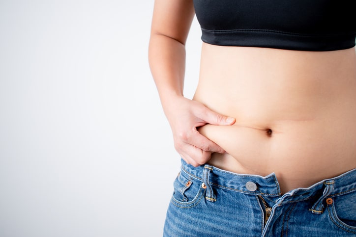 A close up of a woman's hand pinching her own belly fat in jeans and a sports bra