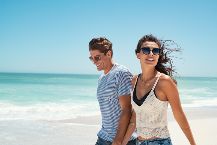 A young couple walks along a sunny beach hand in hand and wearing sunglasses