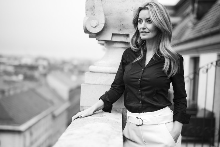 A beautiful older woman with wavy hair and a fancy black blouse looks out from an ornate patio