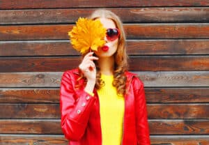 A woman in a yellow sweater, red leather jacket, and heart-shaped sunglasses makes a pouty kiss face and holds a large autumn maple lead over her eye