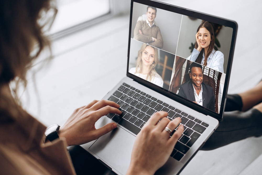 A woman has a laptop on her lap and is looking at a virtual conferencing meeting with 4 other people on the screen