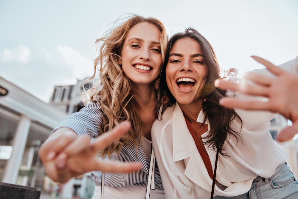 Two female friends laugh and make the peace sign while posing outside