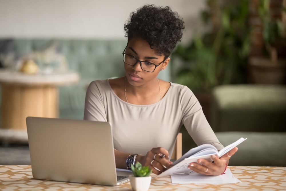 A young woman in glasses and professional clothes looks at a laptop and holds a notebook