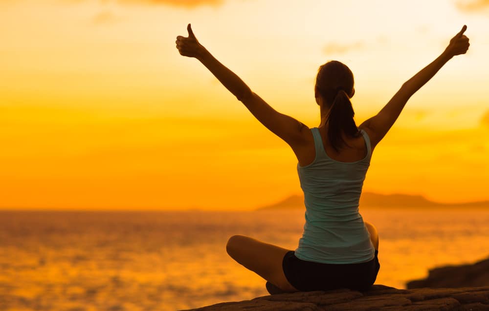 A woman sits cross legged in front of the ocean at sunrise and holds her fists up in the air with two thumbs up